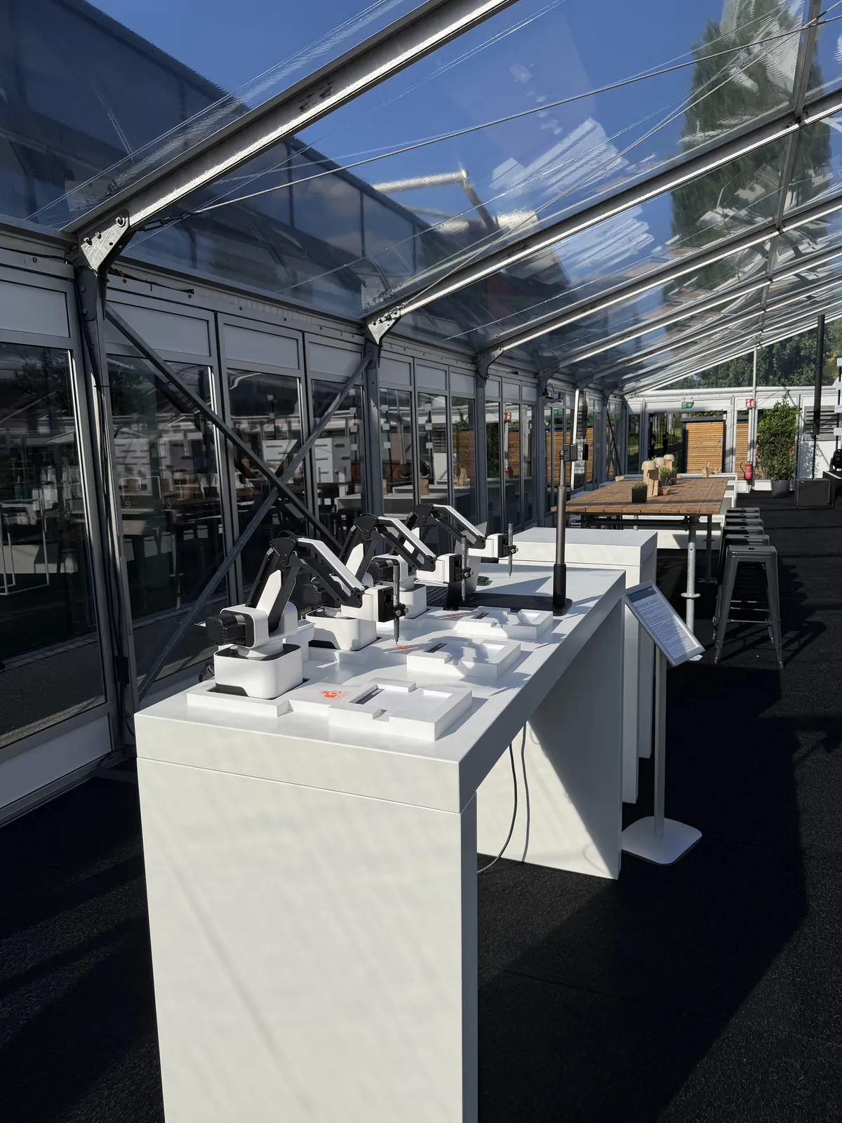 KI Zeichenroboter auf weißen Stehtischen in einem lichtdurchfluteten Glaspavillon auf dem Gelände der Messe Stuttgart